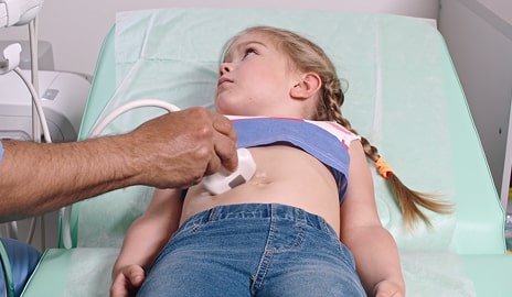 A child lies on an examination table receiving an ultrasound in a hospital room, with medical staff present.
